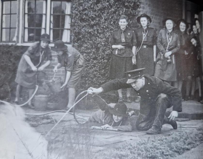 A group of Girl Guides in WW2 uniform operate a stirrup pump in a bucket of water whilst another helps a fireman direct the hose on a fire.